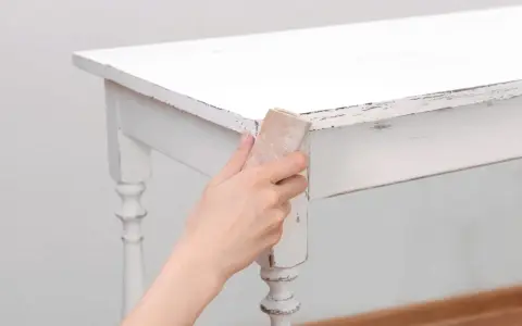 Main tenant, comment décaper un meuble peint. Une main utilise du papier de verre pour poncer le bord d'une table blanche écaillée.