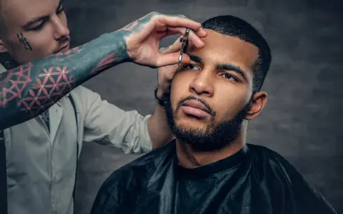 Barbero con tatuajes en el brazo recorta las cejas de un hombre con barba, creando un look buzz cut.