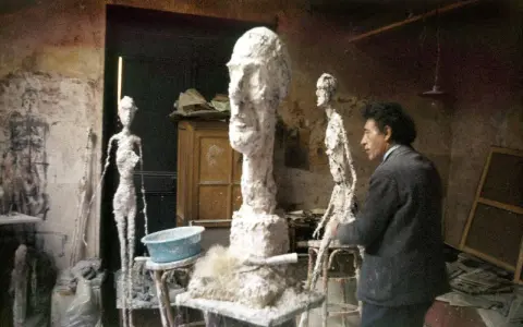 Alberto Giacometti w pracowni, otoczony swoimi charakterystycznymi, smukłymi rzeźbami i niedokończonymi formami.