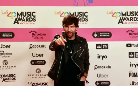 Hombre con chaqueta de cuero negro señala a la cámara en una alfombra roja de Los40 Music Awards. ¡Parece que ha pescado el premio!