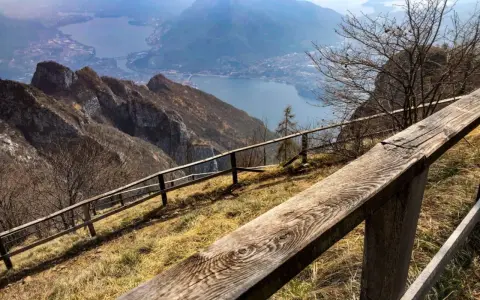 Sentiero con staccionata in legno per una passeggiata sul Pian dei Resinelli, con vista panoramica sul lago e le montagne.