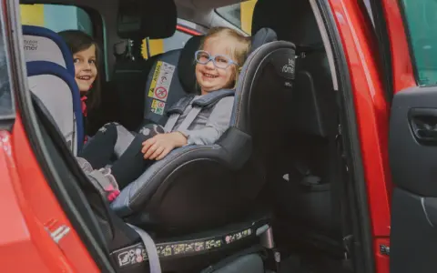 Dwie uśmiechnięte dziewczynki w fotelikach Britax Romer 0-18 kg w czerwonym samochodzie.