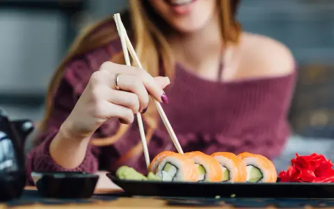 Uśmiechnięta kobieta w ciąży sięga po sushi pałeczkami. Na talerzu łosoś, awokado i imbir.