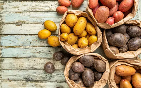 Une variété de pommes de terre, allant du jaune doré au rouge et au violet foncé, est présentée dans des sacs en papier sur une table en bois vieilli.