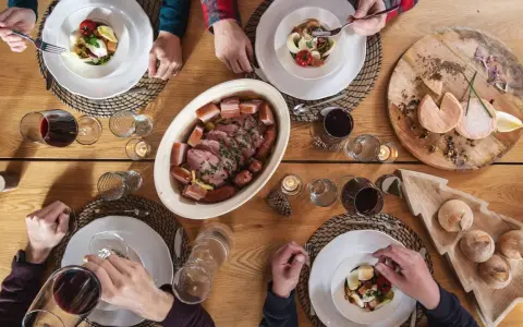 Un repas du terroir avec viande, saucisses, fromages et pain sur une table en bois. Des mains servent et dégustent.
