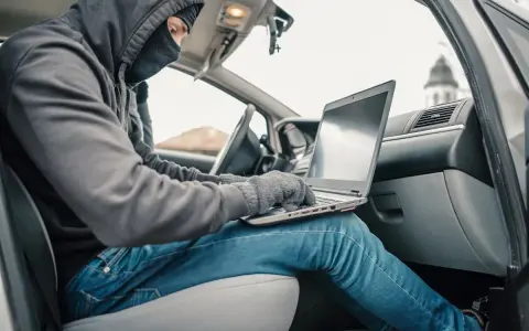 Haker w masce i kapturze próbuje obejść zabezpieczenia keyless go, używając laptopa w samochodzie.