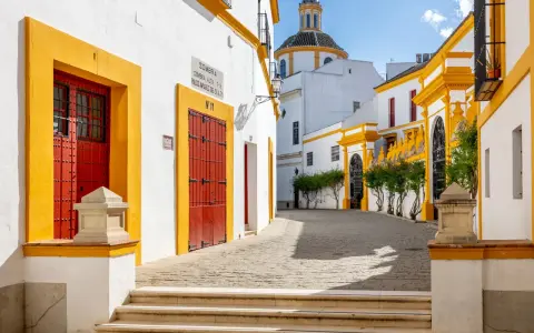 En solig gränd i Sevilla, Spanien, med vita hus, gula detaljer och röda dörrar. En kupol syns i bakgrunden.