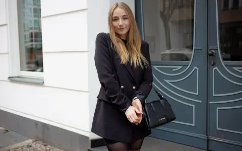 Frau mit blonden Haaren trägt ein schwarzes Kleid mit Blazer, kombiniert mit einer schwarzen Tasche.