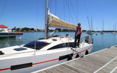 Un homme en gilet rouge ajuste une aussière sur un bateau blanc, prêt à appareiller. D'autres voiliers sont au mouillage.