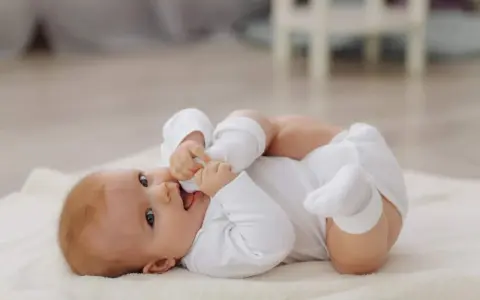 Niemowlę w białym body leży na kocyku, gryząc swoje rączki. Po trzydniówce z wysypką, maluch dochodzi do siebie, gotowy na pierwszy spacer.