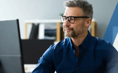 Hombre con gafas y barba gris, concentrado frente a su ordenador. Su camisa azul oscuro es un ejemplo de **art 16 et** en el vestuario profesional.