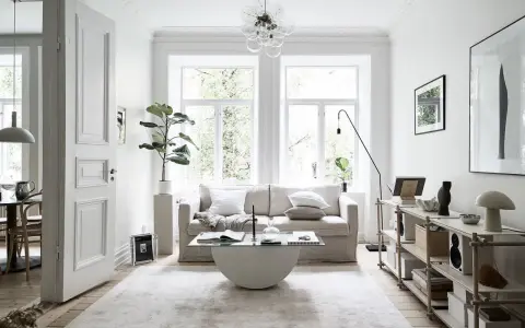 Salón con sofá beige, mesa de centro de cristal y alfombra clara. Decoración minimalista y plantas, creando un ambiente de feng shui.
