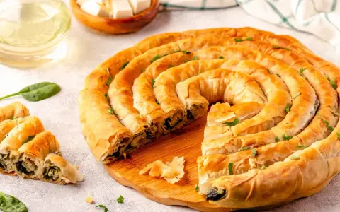 Złocisty borek ze szpinakiem, zwinięty w spiralę, kusi chrupiącym ciastem i aromatycznym nadzieniem. Obok kilka porcji i świeże liście szpinaku.