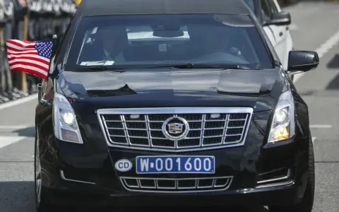 Czarny Cadillac z amerykańską flagą i tablicą rejestracyjną CD W-001600, wskazującą na rejestracje dyplomatyczne.