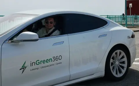 Uśmiechnięty mężczyzna w okularach przeciwsłonecznych siedzi za kierownicą białej Tesli z logo inGreen360.