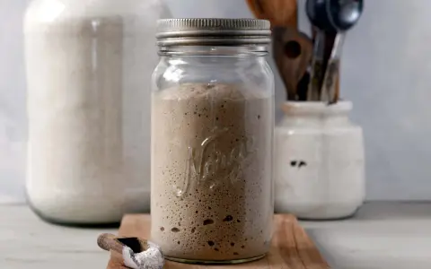 Blubbernde Sauerteig Kultur in einem Glas, bereit für das nächste Brot. Daneben ein Löffel mit Mehl und ein großer Behälter mit weiteren Zutaten.