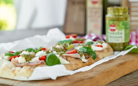 Długi placek z mozzarellą, pomidorkami i bazylią, polany zielonym pesto. Obok słoiczek z pesto.