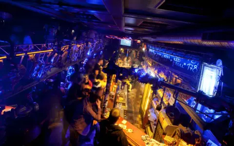 Lebhafte Nacht in einem der Prager Clubs, beleuchtet von blauem und gelbem Licht. Menschen stehen an der Bar und unterhalten sich.