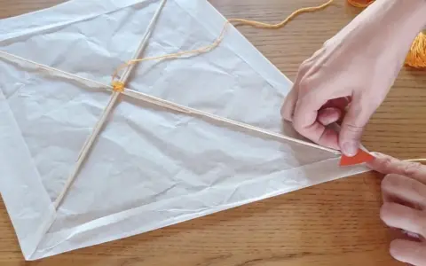 Mains fabriquant un cerf-volant simple en papier blanc et bâtons de bois, avec fil jaune.