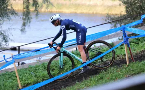 Cyclo cross racer in blue kit navigates a muddy course beside a lake, marked by blue tape.
