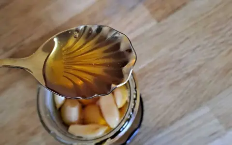 Une cuillère dorée verse du miel ambré sur des gousses d'ail confites dans un bocal. Un délice d'ail au miel.