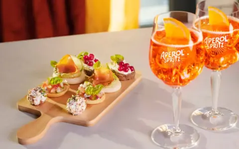 Zwei Aperol Spritz Gläser mit Orangenscheiben und eine Holzplatte mit verschiedenen Häppchen. Perfekt für einen Aperitif!