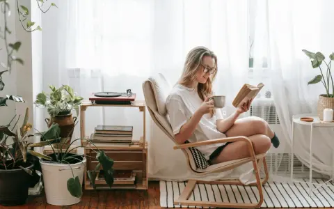 Mujer con gafas disfruta de un libro y una taza en su rincón de lectura, rodeada de plantas y luz natural.