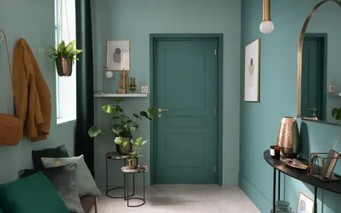 Entrada decorada con pared verde azulado, puerta verde, perchero con abrigo y bolso, plantas y espejo.