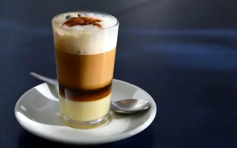 Ein geschichteter Kaffee con leche in einem Glas, mit einer Löffel auf einer Untertasse.