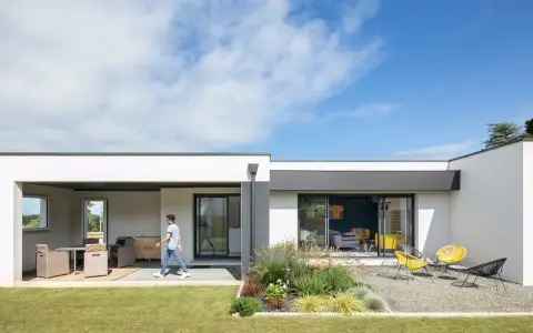 Une jeune personne traverse la pelouse devant une petite maison moderne avec une terrasse aménagée et un jardin de gravier.