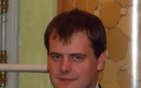 Tomasz Bartoś, zamyślony, w eleganckim stroju, na tle ozdobnej ściany.
