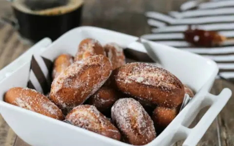 Beignets alsaciens schenkele dorés et saupoudrés de sucre, servis dans un plat blanc avec une tasse de café en arrière-plan.