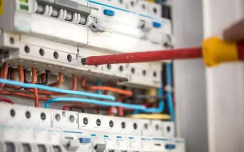 Ręka z czerwonym śrubokrętem pracuje przy instalacji niskoprądowej. Widać bezpieczniki i kolorowe przewody.