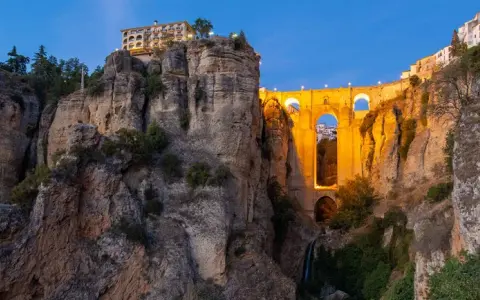 A dramatic **Ronda bridge view point** at dusk, with the illuminated bridge spanning a deep gorge and buildings perched on the cliff edge.