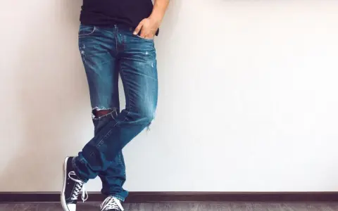 Mężczyzna w modnych jeansach z dziurami na kolanie, czarnych trampkach i czarnej koszulce, opiera się o ścianę.