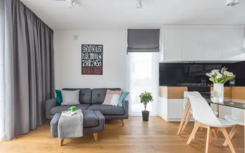 Mały salon inspiracje: szara kanapa, stolik z pufą, nowoczesna kuchnia i jadalnia. Drewniana podłoga dodaje ciepła.