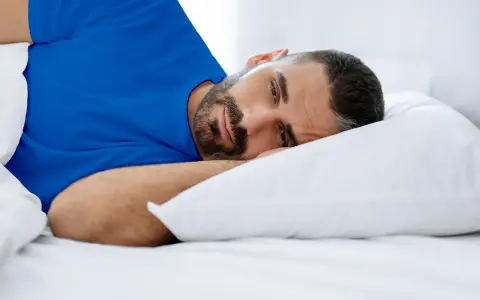 Hombre con barba en cama, con ojos abiertos, vistiendo camiseta azul. Duermo pero no descanso, ¿qué puedo hacer?