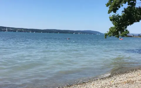 Sonniger Bodensee-Strand, ein echter Geheimtipp. Segelboote gleiten über das Wasser, während ein paar Schwimmer die Ruhe genießen.