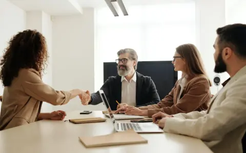 Uśmiechnięty mężczyzna ściska dłoń kandydatce, budując pozytywny employer branding. Przy stole siedzą też inne osoby.