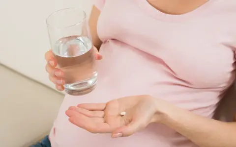 Kobieta w ciąży trzyma tabletkę i szklankę wody, szukając odpowiedzi na pytanie: co na ból głowy w ciąży.