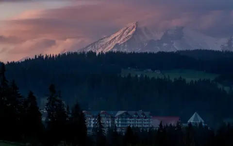 Górski krajobraz o zachodzie słońca z ośnieżonymi szczytami, lasem i budynkami hotelowymi.