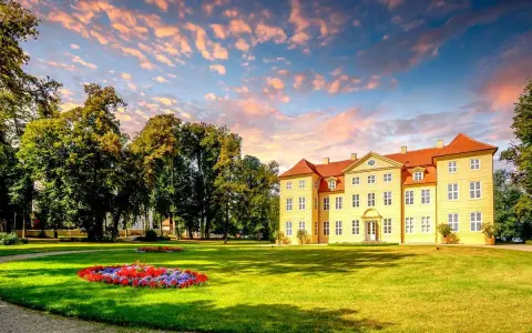 Gelbes Schloss mit roten Dächern, umgeben von grünen Bäumen und Blumenbeeten. Ein malerischer Anblick auf der Liebesinsel Mirow.