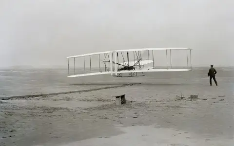 Der Wright Flyer hebt vom sandigen Boden ab, ein Mann beobachtet den historischen Flug.