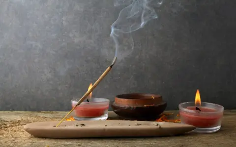 Un incienso humeante se posa sobre un soporte de arcilla, rodeado de velas rojas y un cuenco de madera. Perfectos inciensos para limpiar energías y crear un ambiente de paz.
