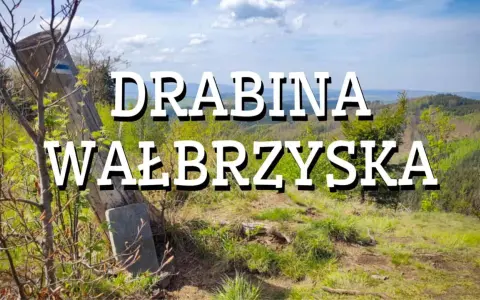 Widok na górskie krajobrazy z napisem "Drabina Wałbrzyska". Stary drewniany słup z niebieskim znakiem szlaku stoi na zboczu porośniętym trawą i młodymi drzewkami.