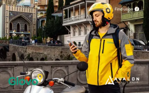 Kurier Glovo sprawdza telefon, gotowy do pracy w blokach czasowych. Na skuterze widnieje logo.