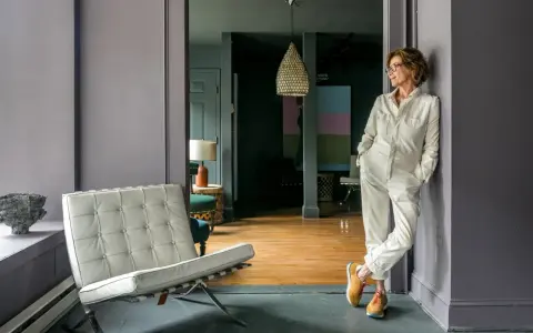 A woman in a cream jumpsuit leans against a wall in the robin rice gallery, with a Barcelona chair and abstract art visible.