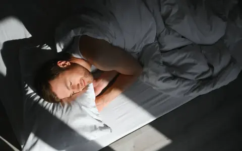 Mężczyzna śpi, otulony kołdrą. Promień słońca pada na jego twarz, podkreślając spokój osoby wysoko wrażliwej.