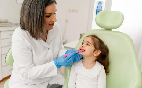 Dentystka w białym fartuchu i niebieskich rękawiczkach pokazuje małej dziewczynce, jak prawidłowo szczotkować zęby. To przykład troski o zdrowie jamy ustnej w ramach pedodoncji.