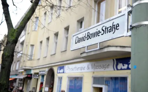 Straßenschild "David-Bowie-Straße" in Berlin. Ein Baum und ein Gebäude mit Geschäften im Hintergrund.